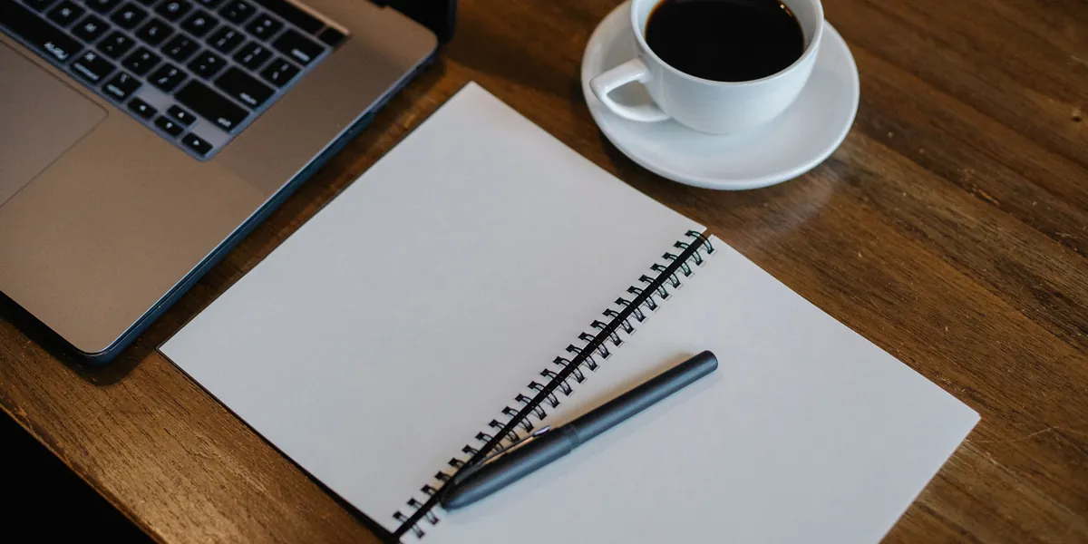 An open, blank notebook with a pen on it, a cup of coffee and the edge of a laptop on a dark wooden table, seen from above