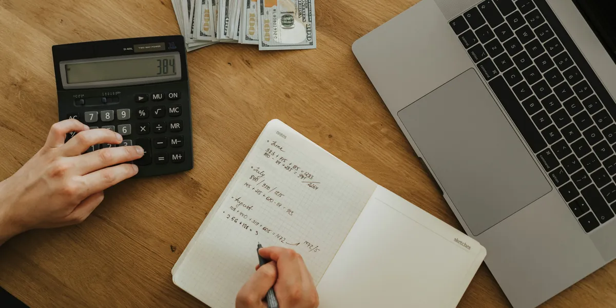 Hands typing on a black calculator next to a notebook with handwritten monthly sums, a laptop and a few banknotes, seen from above