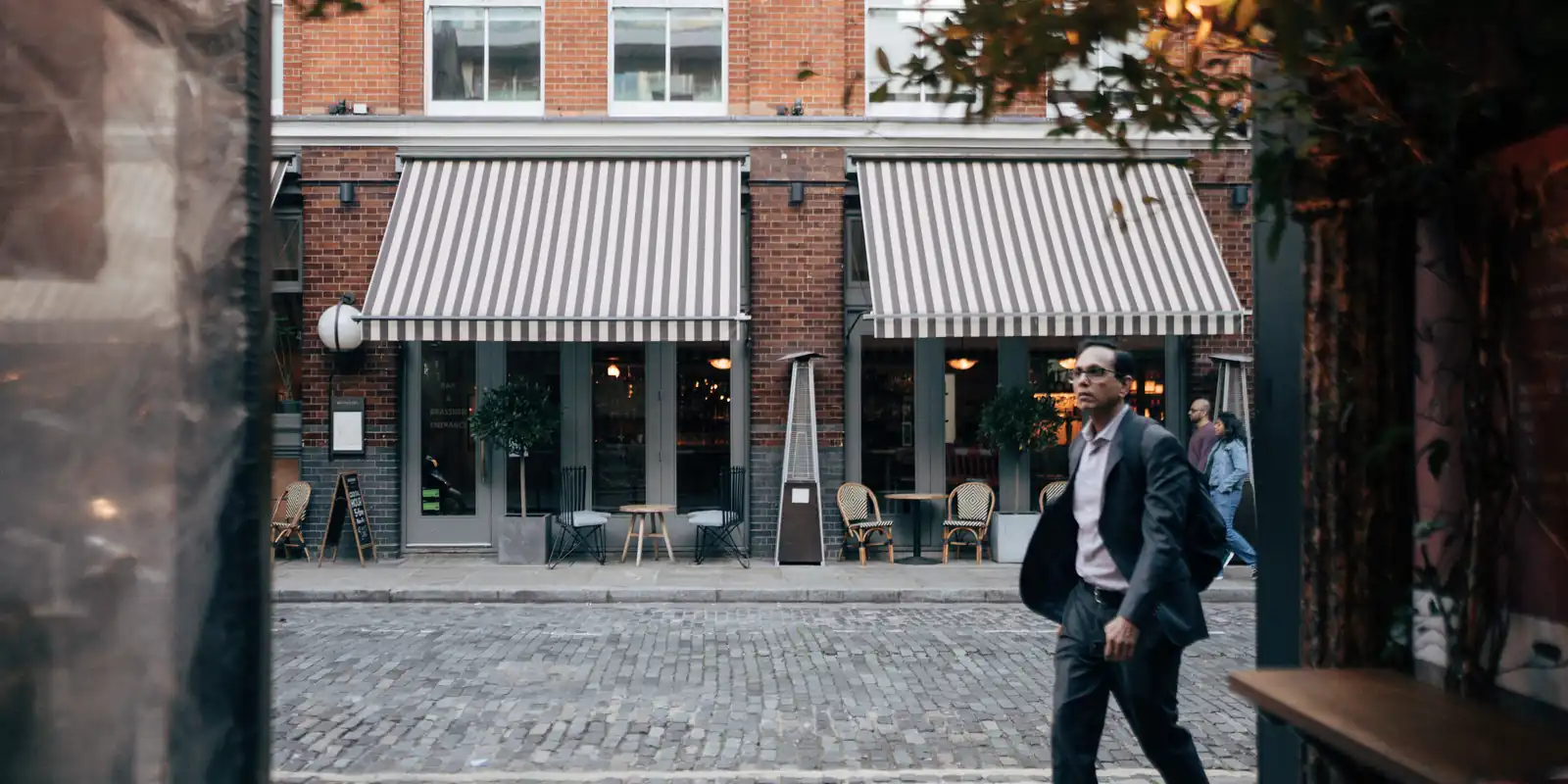 The terrace of a café with a striped awning on a cobbled street, a man in a suit walking past, towards evening