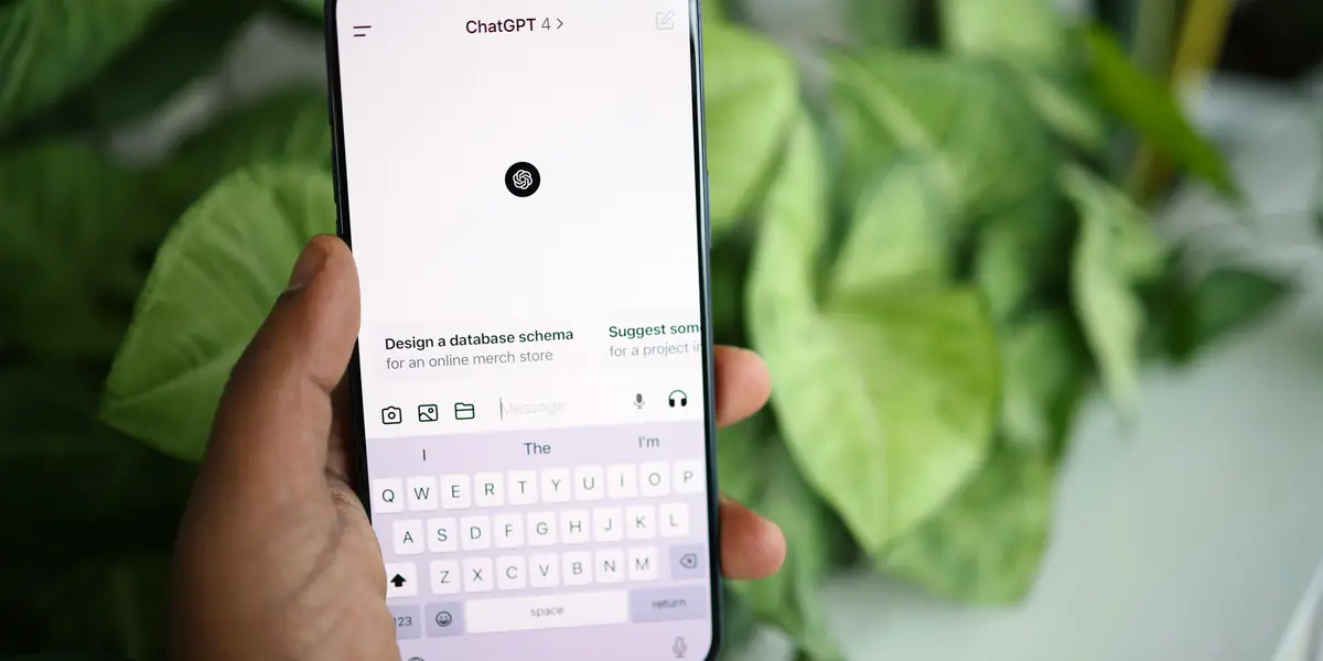 A hand holding a phone with the ChatGPT 4 app open, the on-screen keyboard raised and the message box empty, with the suggestion card Design a database schema for an online merch store above it, a green houseplant blurred behind