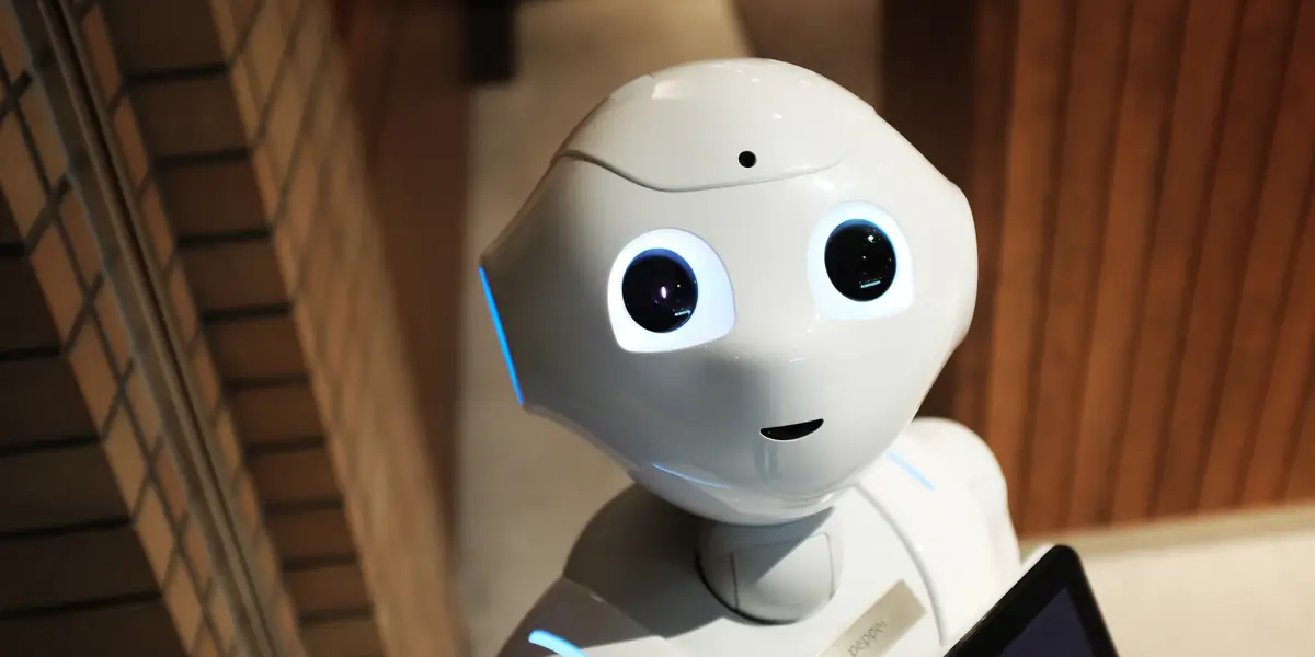Robot umanoide bianco con grandi occhi neri rotondi e targhetta con la scritta pepper sul petto, davanti a una parete di legno