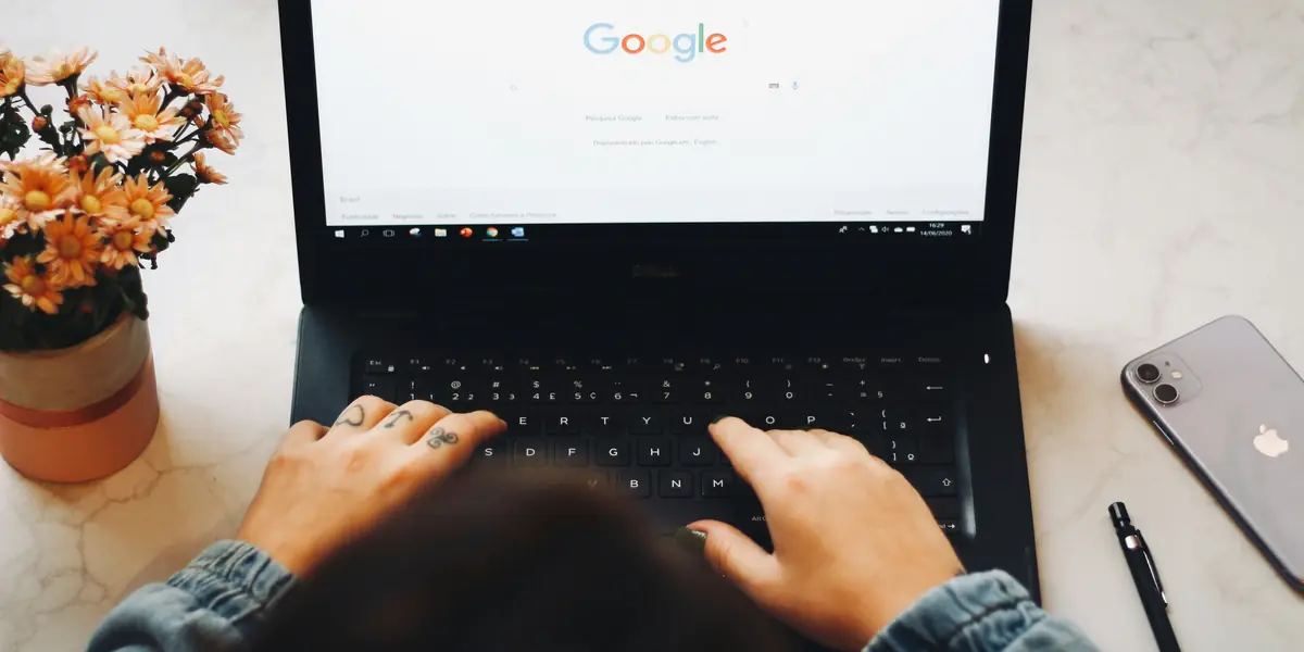 Overhead view of a marble desk: two hands typing on an open laptop showing the Google home page, with a silver phone, a black pen and a pot of orange flowers around it