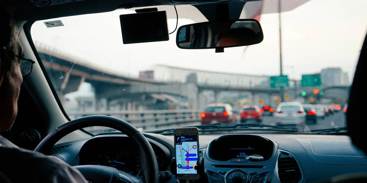 Vedere din scaunul șoferului: telefon prins de bord cu o hartă de navigație pe ecran, trafic dens în față și un pasaj rutier peste bandă