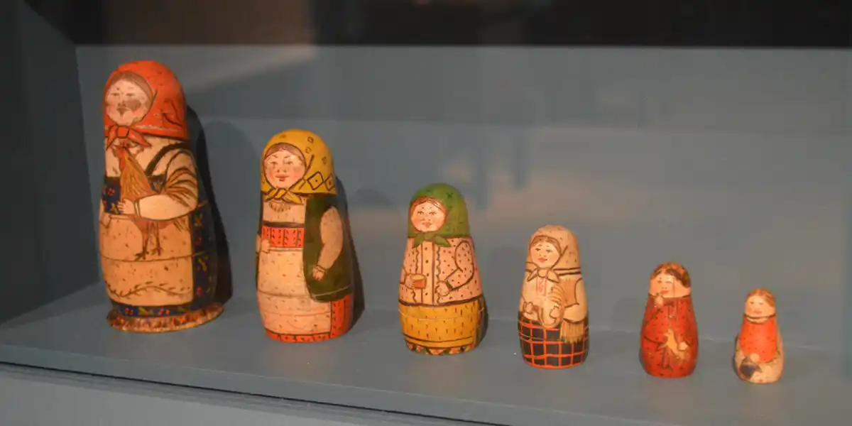 Row of six antique wooden nesting dolls in a museum display case, arranged from tallest to smallest, each painted with a different folk figure