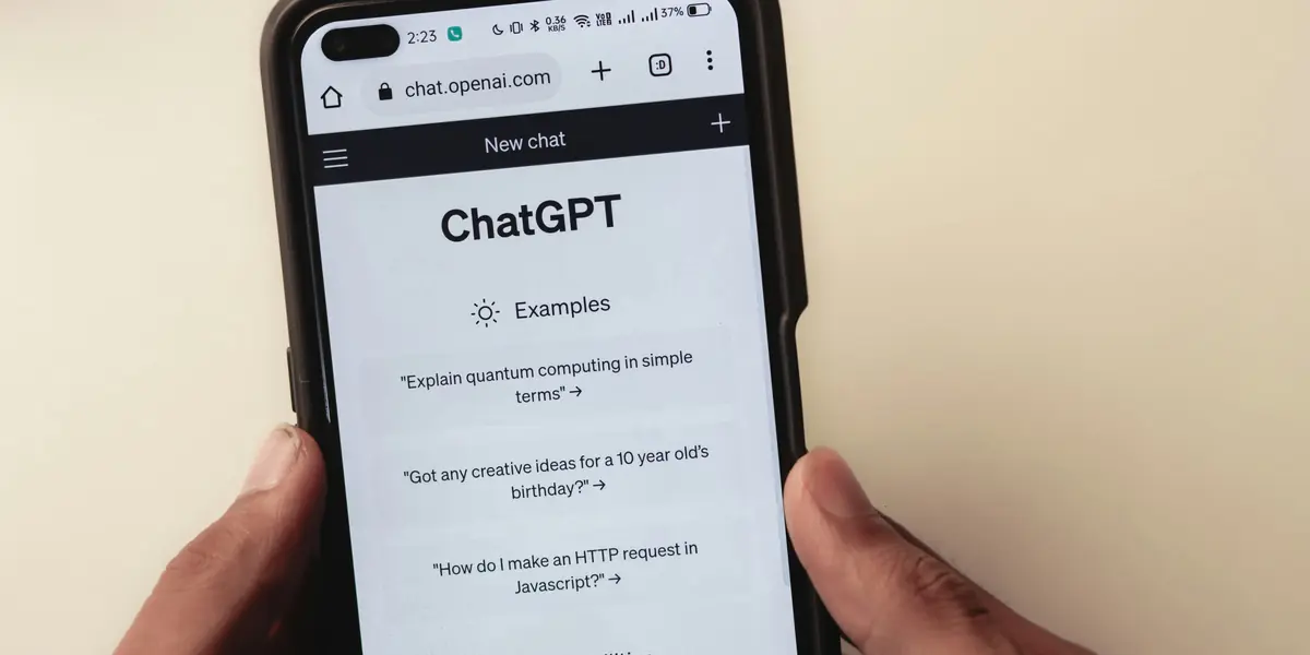 Două mâini țin un telefon negru pe care e deschis ChatGPT într-un browser, la ecranul de chat nou, cu trei exemple de întrebări listate dedesubt și indicatorul de baterie la 37 la sută
