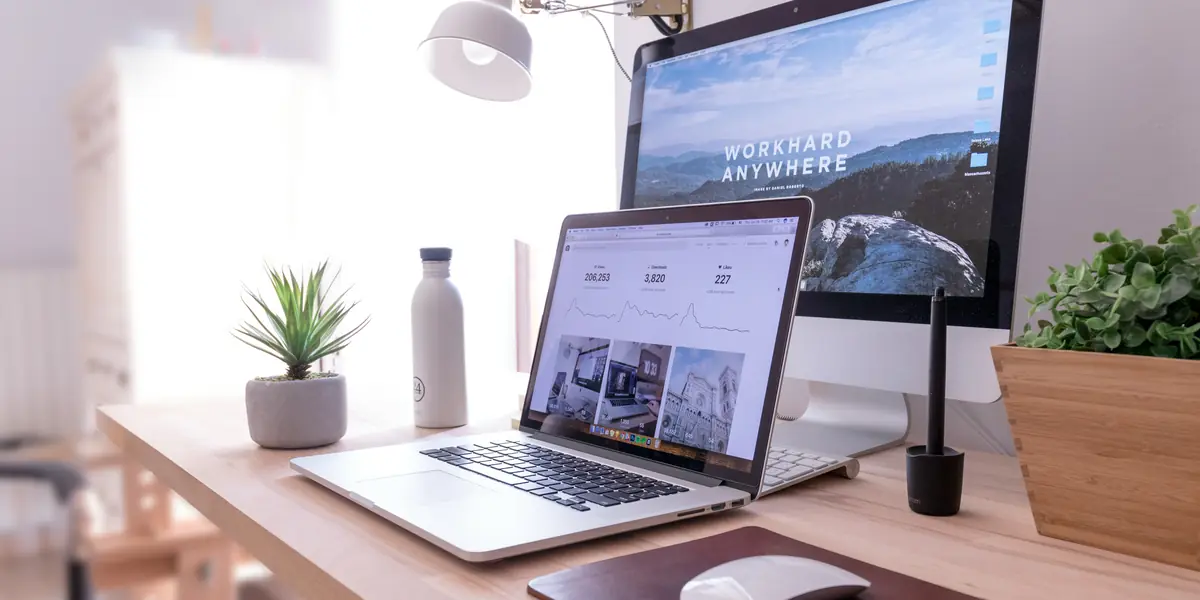 Bright desk with an iMac showing the words Work hard anywhere over a mountain photograph, in front of it an open laptop displaying an analytics panel with the figures 206,193, 3,820 and 227 above three charts, next to a white metal bottle and a small succulent